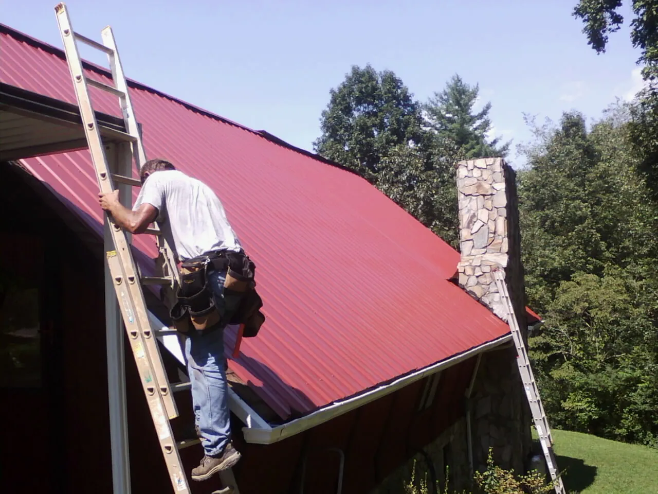 Roofer on a ladder beside a red metal roof and stone chimney — Soffit & Fascia Repair by Roofing Brothers in Shepherdstown